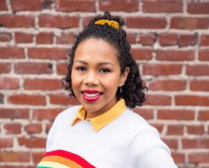 A Black person with shoulder-length black hair pulled back with a yellow scrunchie, red lipstick. They are smiling and wearing a white sweater with a yellow collar, which has a rainbow across the middle of it. They're against a brick wall.