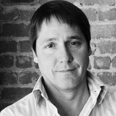 A black and white photo of Allen Hopper in front of a brick wall, wearing an open button down striped shirt.
