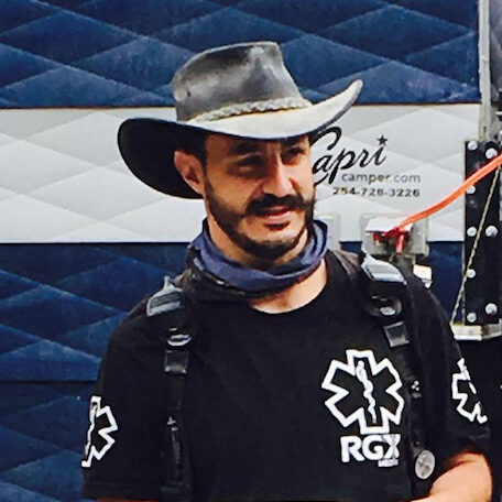 A photo of Richard standing in front of a camper wearing a cowboy hat, radios, and a black RGX Medical shirt.