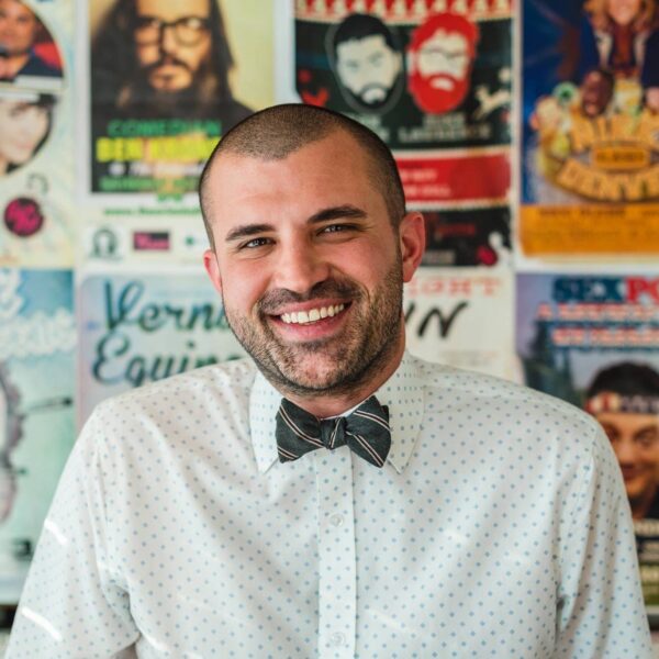 A photo of Kayvan smiling at the camera wearing a polka dotted white button down shirt with a green bow tie, in front of a wall of posters.