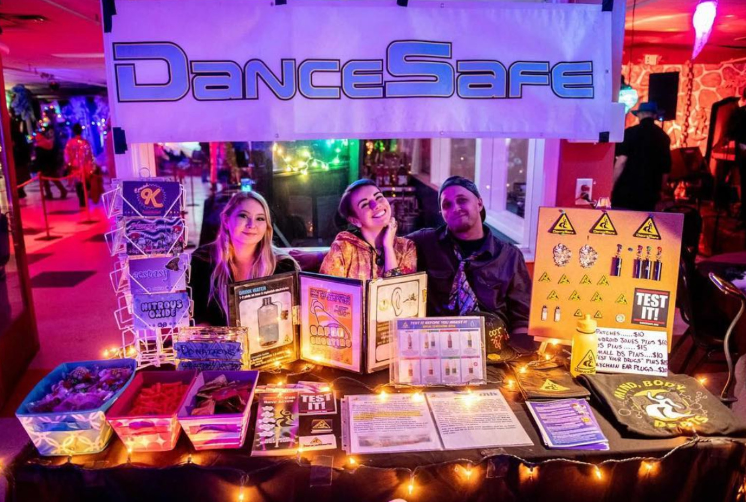 Three people sit behind a table, all with light-toned skin. The table is softly lit by warm orange lights and contains bins of condoms, earplugs, and other harm reduction supplies, neatly organized next to pamphlets and pegboards of pins and earrings. A white poster with the DanceSafe logo in blue hangs above and behind the three people, who have, from left to right, long blonde hair and a black sweater on, brown hair and an orange and red hoodie on, and a short beard and a brown jacket on. Glowing red and blue lights of a party are visible in the background.