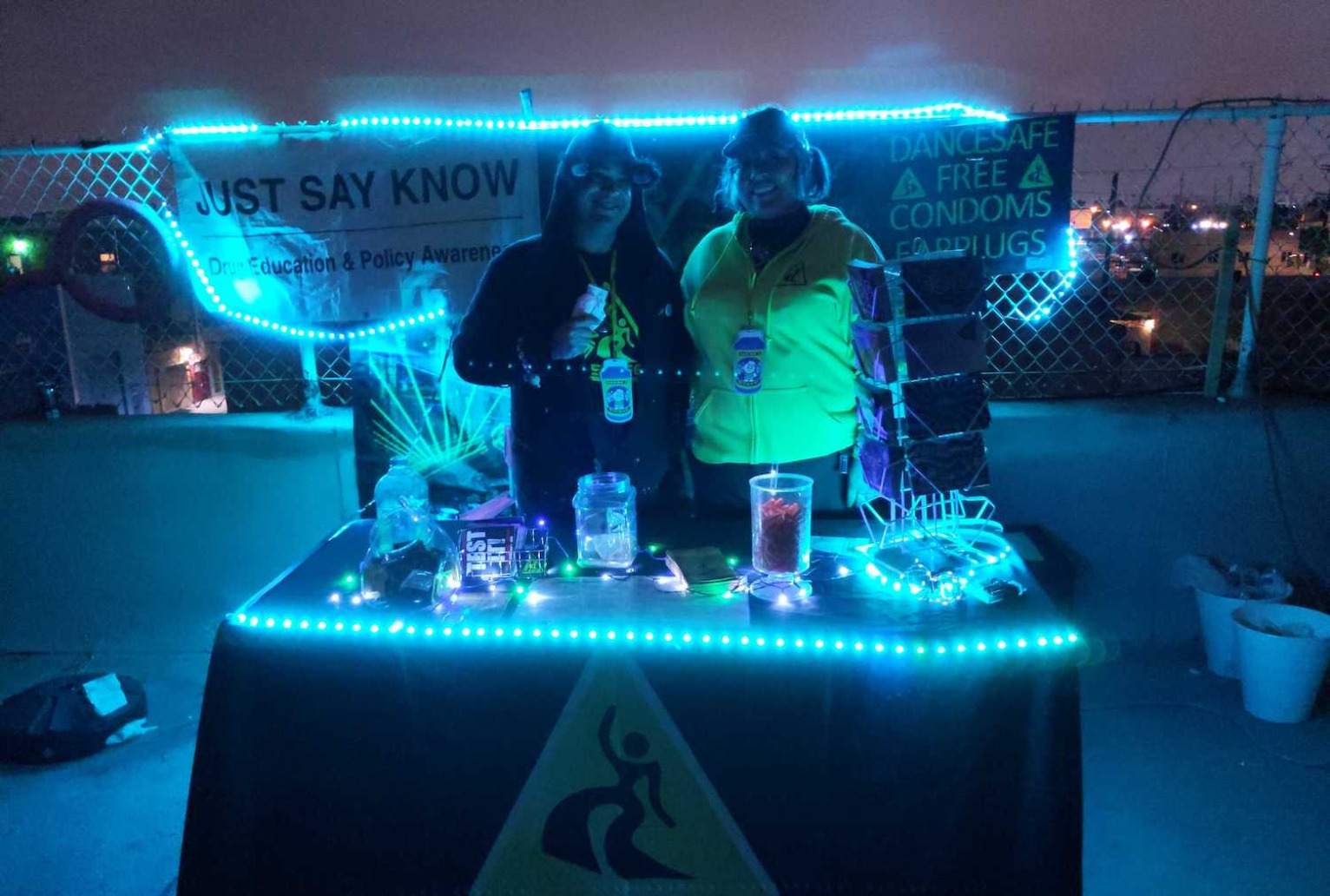 Two people, backlit with faces not very visible, stand behind a small table with a black DanceSafe-branded tablecloth on it. The table and the fence behind it are lined with glowing blue LED lights, which illuminate harm reduction supplies like earplugs and condoms scattered across the table. The people are wearing, respectively, a black sweater with the DanceSafe logo on it and fuzzy ears, and a bright yellow shirt with the DanceSafe logo on it and a hat. Behind them are posters saying things like "just say know" and "free condoms" hung up on a chain link fence. They appear to be on the top of a building, with a concrete floor and a concrete segment behind them below the fence.