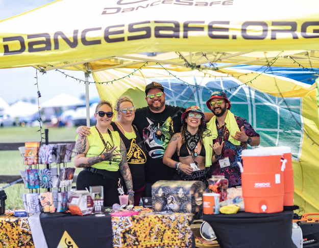 Five people stand close together behind a table covered in a black DanceSafe-branded tablecloth and a variety of harm reduction supplies. There's a yellow DanceSafe-branded canopy above them, with windowpane outlines on the back wall, and two orange 5-gallon jugs on the right side of the table. The people are all smiling, wearing a mixture of black and yellow DanceSafe-branded clothes for the most part, all lighter-toned skin and all wearing sunglasses. White festival tents are visible in the distance.