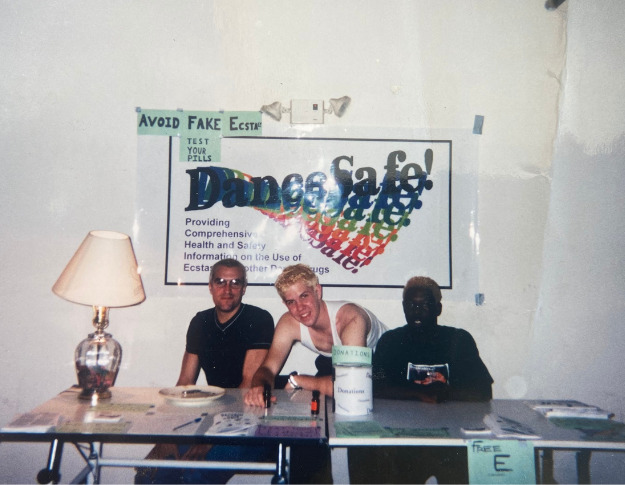 Three people sit behind a table. It's clear that the photo is old (it's from 1999!), and there's a white poster on a white wall that reads "DanceSafe!" in navy blue letters with a rainbow of letters spilling out underneath "safe." Barely legible text written on blue paper above the sign reads "Avoid fake ecstasy." A cream-colored lamp sits on the left hand side of the table. The leftmost person has light-toned skin and a buzz cut, wearing a black shirt and reflective sunglasses. Next to him is a person with light-toned skin and short blonde hair, wearing a white tank top and leaning over the middle of the table, smiling. To their right is a person with dark-toned skin and short, curly orange-toned hair, wearing a black shirt with an illegible graphic on it. Various papers and supplies are scattered across the table they're sitting at.