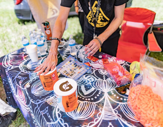 The shot is a slanted aerial view of a blue and white patterned tablecloth on a table of harm reduction supplies. Two orange containers of Gatorade are in the foreground, one of which has a light-toned hand pressing down on its lid. The person whose hand it is is barely visible wearing a black and yellow DanceSafe shirt. Various other harm reduction supplies like orange earplugs and water bottles are scattered across the table, and you can see green grass on the other side of it as well as a red lawn chair behind the person.