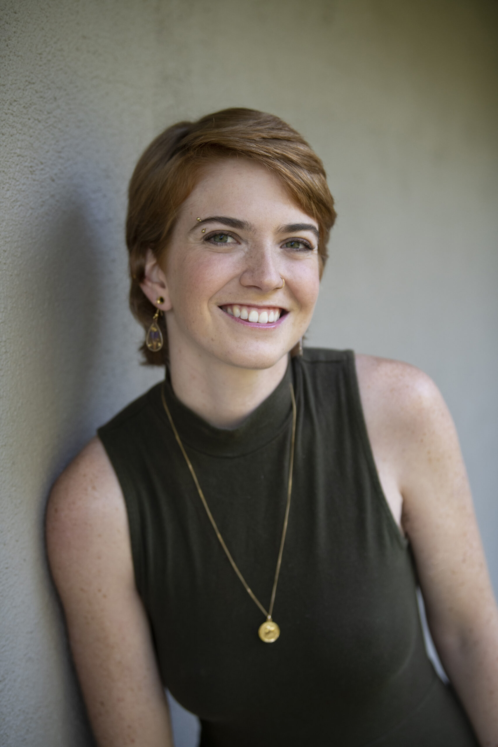 Partial-body headshot of Rachel leaning against a grey wall in a no-sleeve olive green dress.