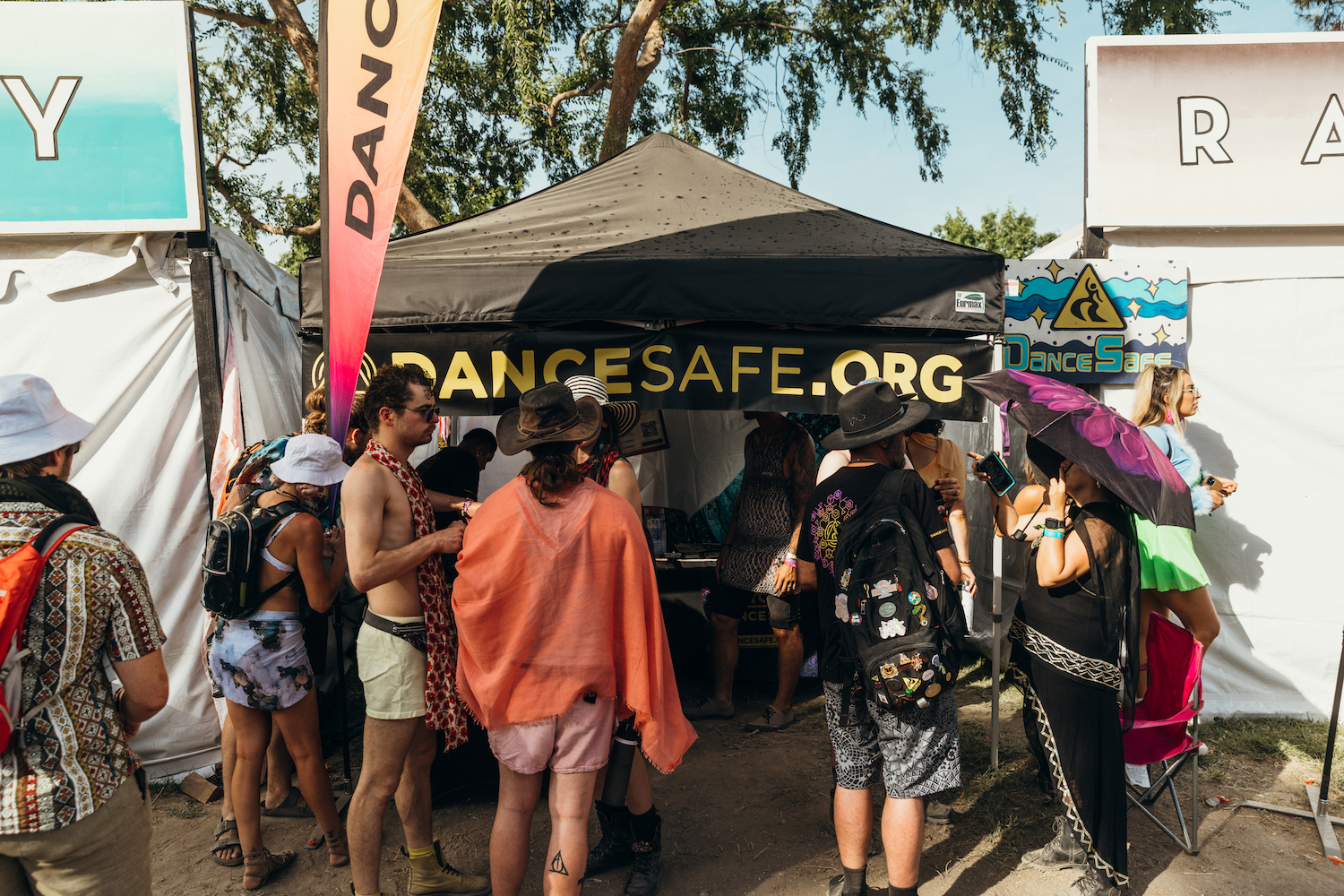 A group of people stand in front of a DanceSafe booth with a black canopy, not looking at the camera. They have all kinds of different appearances. A DanceSafe flag is visible to the left of the booth, a soft pink to peach ombre, and on either side of the booth are two large white structures that have lettering peeking out (but not legible). There's a large tree overhead, a blue sky in the background, and dirt on the ground.