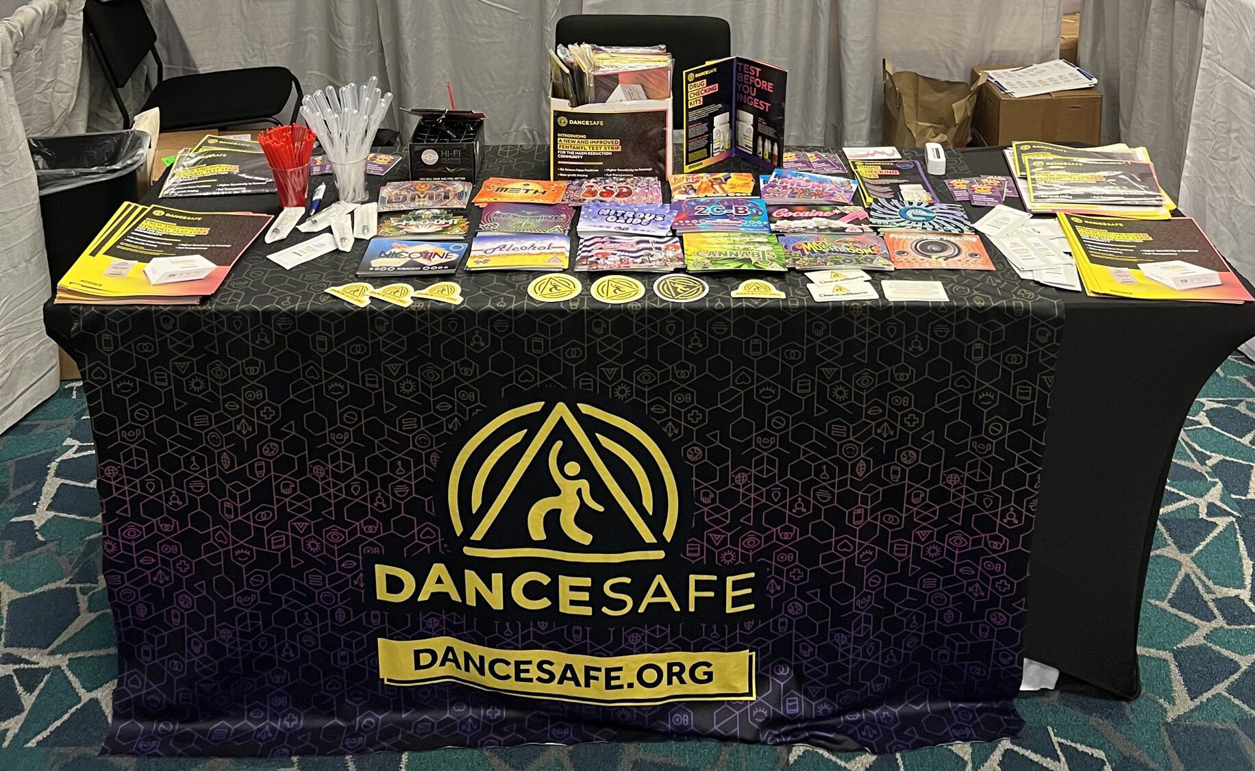 A photo of the DanceSafe booth at a conference, covered in colorful drug info cards, stickers, and fentanyl test strips.