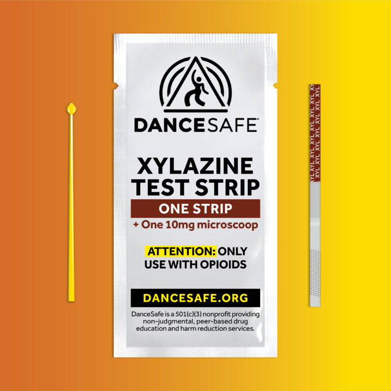 DanceSafe Xylazine Test Strip package displayed against an orange-to-yellow gradient background. The white packet features the DanceSafe logo and reads “Xylazine Test Strip – One Strip + One 10mg Microscoop.” A yellow microscoop and a single test strip are shown beside it. The label includes a warning in yellow and black text: “ATTENTION: Only use with opioids,” and the website “DanceSafe.org.